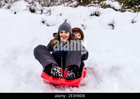 two very happy caucasian kids playing in the snow on a mountain with a red sled Stock Photo