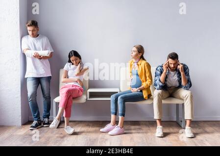 Multiethnic people waiting in queue near pregnant woman and man with gypsum o hand in clinic Stock Photo