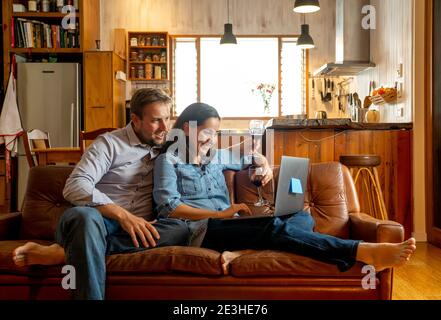 Life at home image of Happy couple video calling friends and family using laptop. Man and woman online chatting cheering with wine in virtual celebrat Stock Photo