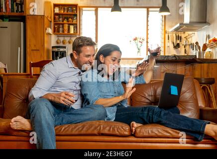 Life at home image of Happy couple video calling friends and family using laptop. Man and woman online chatting cheering with wine in virtual celebrat Stock Photo