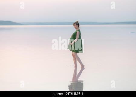 Pregnant woman in green dress posing on the beach. Young happy pregnant woman relaxing and enjoying life in nature Stock Photo
