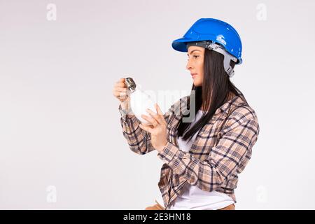 A female electrician in a blue hard hat examines an LED lamp on a white background with empty side space. Stock Photo