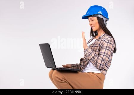 New norms and online concept. Woman in hard hat greeted by video communication while holding laptop on her feet. White background. Stock Photo
