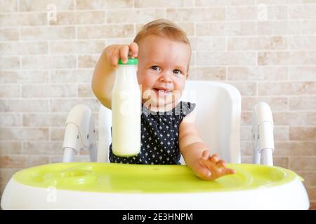 Pretty baby girl holding bottle with milk. Kid sitting in the chair at home. Smiling child is 7 months old Stock Photo