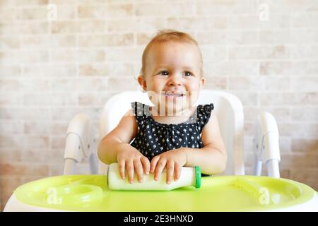 Pretty baby girl holding bottle with milk. Kid sitting in the chair at home. Smiling child is 7 months old Stock Photo