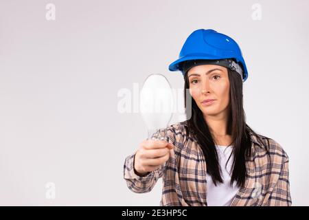 A serious woman in a blue hard hat holds out a front hand towards the camera LED lamp on a white background with empty side space. Stock Photo