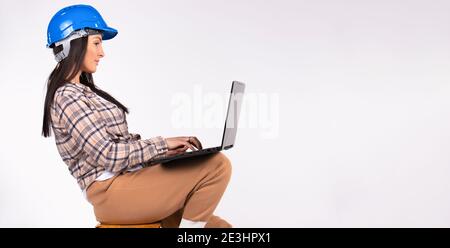 Banner. A woman sits on a stool in a blue hard hat and holds a laptop on her feet. White background and empty side space. Stock Photo
