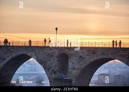 People walking on a stone bridge at sunset and the frozen trees in winter at the golden hour. Stone arches, reflection of the sun on the river. Two br Stock Photo