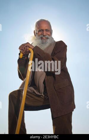 A BEARDED OLD MAN LOOKING AT CAMERA AND HAPPILY POSING WITH WALKING STICK Stock Photo