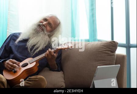 A BEARDED OLD MAN HAPPILY SITTING AND PLAYING UKULELE INSTRUMENT Stock Photo