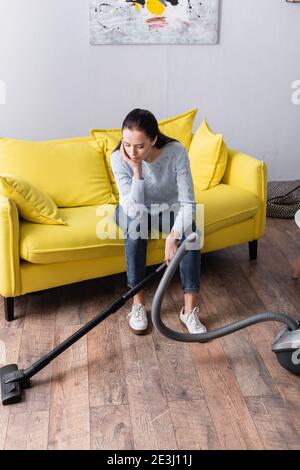tired housewife sitting on yellow sofa near vacuum cleaner Stock Photo