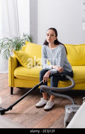 upset, tired housewife sitting on couch near vacuum cleaner Stock Photo