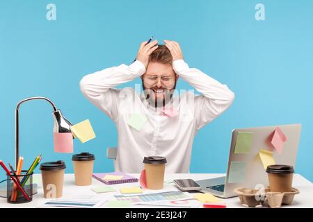 Scared busy man analyst with beard sitting at workplace with coffee cups all covered with sticky notes, has lot of paper work, overworking. Indoor stu Stock Photo