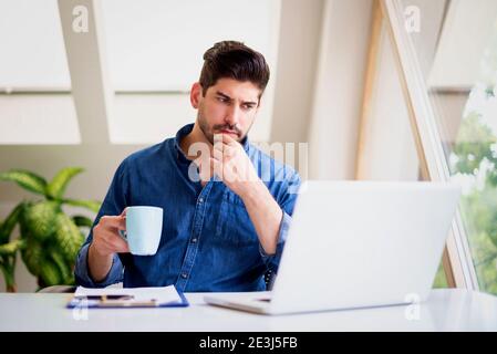 Businessman looking thoughtfully while drinking tea and working on laptop at home. Home office. Stock Photo