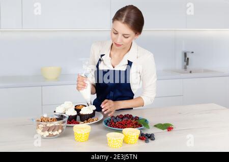 Cute female baker in blue apron  decorating tasty cupcake with creamy top at kitchen table Stock Photo