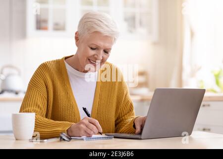 Elderly woman sitting in front of laptop, taking notes Stock Photo