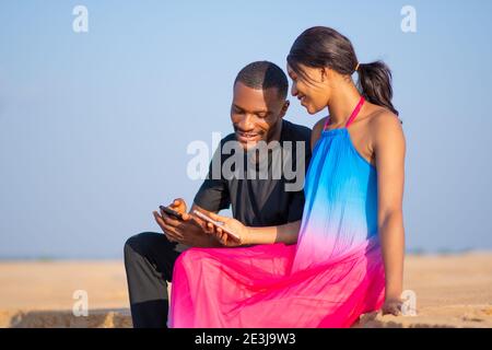 young african man and woman viewing content on a phone together outdoor on a beach, sitting on the sand Stock Photo