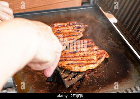 Two pork cutlet steak grilled on electric grill, blurred woman hand holding plastic spatula, about to turn one piece of meat Stock Photo