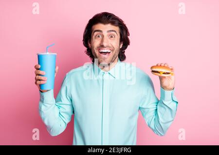 Photo portrait of excited guy holding soda and cheeseburger isolated on pastel pink colored background Stock Photo