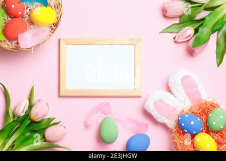 Easter eggs, bunny ears, tulips and frame with copy space on a pink background. Flat lay greeting card. Stock Photo