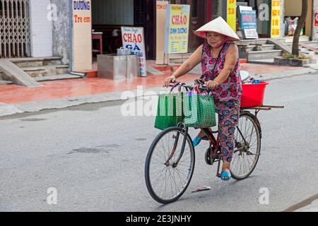 Vietnamese woman riding on bicycle, wearing traditional nón lá, Asian conical hat, in the city Ninh Binh in the Red River Delta of northern Vietnam Stock Photo