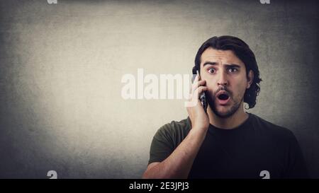 Shocked young man, during a mobile phone conversation, staring with big eyes and mouth open isolated on grey wall background, with copy space. Amazed Stock Photo