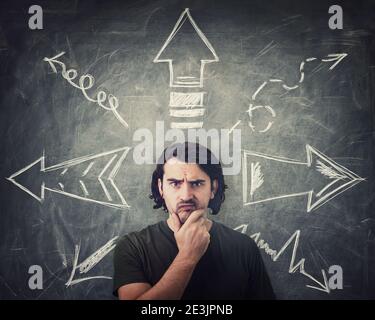 Skeptical guy thoughtful looking to camera, keeps hand under chin, puzzled suspicious expression, has to choose a direction from different arrows poin Stock Photo