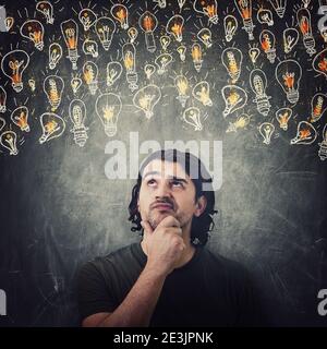 Pensive man, keeps hand under chin, thoughtful gesture, looking up suspicious at different light bulbs sketches. Puzzled and confused casual guy, has Stock Photo