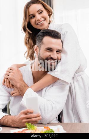 Cheerful woman in bathrobe hugging boyfriend near food on blurred foreground in hotel Stock Photo