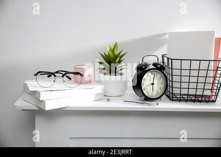A stack of books, an artificial succulent plant in a ceramic pot, glasses, a metal basket with notebooks, a pen and a cup on a white table surface. Co Stock Photo
