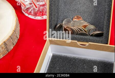 Empty, unwrapped gift box filled with pennies, nickels, dimes, quarters, and a dollar coin, on a red table top with candy canes in the background Stock Photo