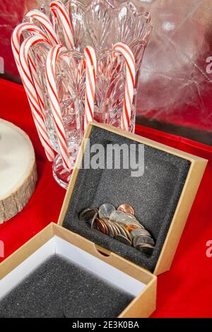 Empty, unwrapped gift box filled with pennies, nickels, dimes, quarters, and a dollar coin, on a red table top with candy canes in the background Stock Photo