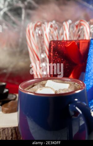 https://l450v.alamy.com/450v/2e3k0ff/blue-and-red-mug-filled-with-hot-chocolate-and-two-marshmallows-with-chocolates-on-a-wooden-stump-and-candy-canes-and-red-and-blue-presents-in-the-ba-2e3k0ff.jpg