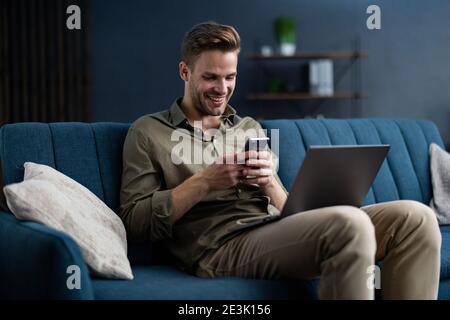 Young freelancer using smartphone and smiling. Happy man using mobile phone apps, texting message, browsing internet, looking at smartphone, sitting Stock Photo