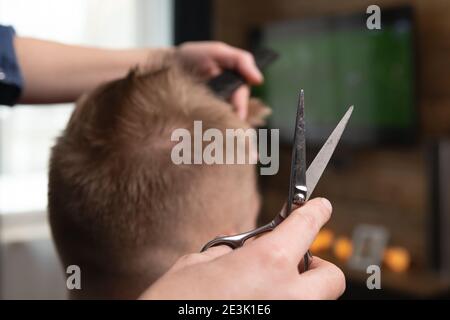 Wife cutting husband's hair at home in front of TV . Scissors in focus. Stock Photo