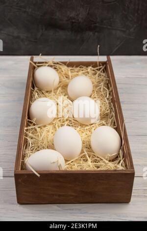 Seven white chicken eggs are lying in a wooden box on the straw. Perspective view Stock Photo