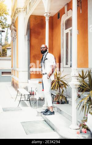 A dashing mature bearded bald black guy in an elegant white suit with a necktie and short sleeve blazer is standing outdoors on the porch of an antiqu Stock Photo