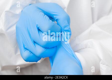 Doctor or nurse in protective PPE suit putting on rubber blue gloves before work with coronavirus patient or test samples or vaccination Stock Photo