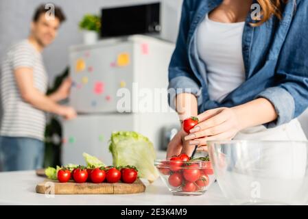 cropped view of woman holding red cherry tomato near ingredients on kitchen table and boyfriend on blurred background Stock Photo