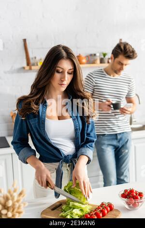 woman cutting lettuce near ingredients on kitchen table and boyfriend on blurred background Stock Photo