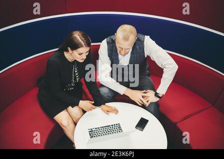 Two business colleagues having a group video call, businessman and businesswoman are sitting on a red curved sofa in an office coworking soundproof ce Stock Photo
