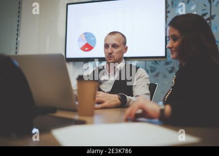 A business conference in a boardroom with two colleagues businessman and businesswoman having a video call using a laptop with partners; plasma tv wit Stock Photo