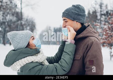 Woman puts mask on boyfriend's face in snowy winter park. Couple in love walking outdoors. Coronavirus covid pandemic safety measures Stock Photo