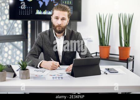 Handsome young bearded businessman in formal wear, posing to camera while sitting at his workplace in modern office, working on digital tablet pc and Stock Photo