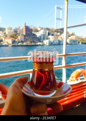 Traditional black Turkish tea in a glass on a plate in the hands with a view of the Bosporus strait and Istanbul background. Travel to Turkey. Stock Photo