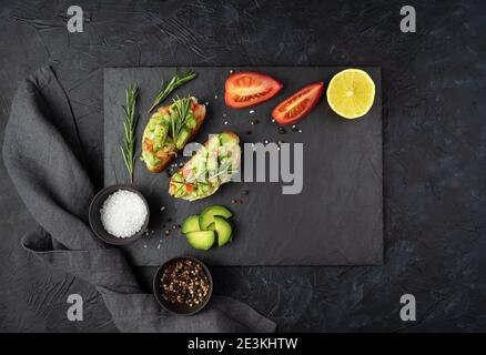 Two avocado sandwiches, tomatoes, lemon and rosemary on a flat plate on a black concrete background. Stock Photo