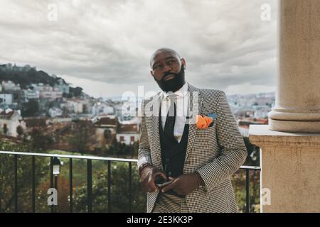 A portrait of a stately mature bearded black guy in a plaid elegant costume with a necktie, he is leaning against a metal fence on the street, on high Stock Photo