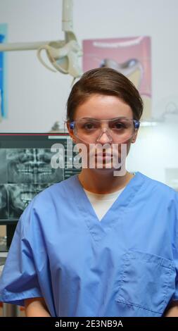 Dental assistant explaining to patients the importance of oral hygiene looking at camera in dental center while doctor is working with patient in background. Stomatologist nurse speaking on webcam Stock Photo