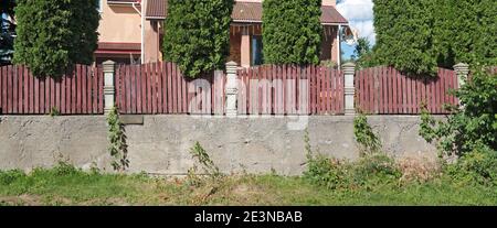 High rustic fence made of concrete and wood. Coniferous trees grow behind the fence. Stock Photo