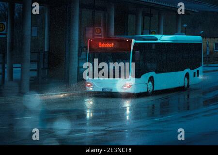 A bus drives to a stop at a bus station in the evening. It is raining. German text means train station. Turquoise colors with complementary color oran Stock Photo